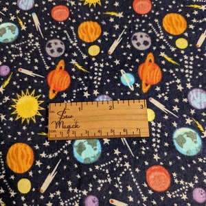 May include: A navy blue fabric with a repeating pattern of planets, stars, and comets. The fabric is measured with a ruler that reads "Sew Magick" in white lettering.