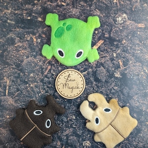 May include: A selection of handmade felt insect and frog appliques. The frog is bright green with white and black eyes. Two beetle-shaped appliques are brown and tan, with white and black eyes. A wooden tag reads "Sew Magick".