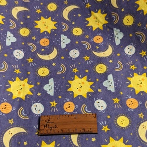Puede incluir: Tela morada con un patrón repetitivo de soles amarillos, planetas naranjas, estrellas blancas y lunas amarillas. Los soles, planetas y lunas tienen caras sonrientes. Una regla se incluye en la imagen para la escala.