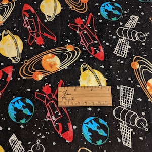 Puede incluir: Tela negra con un estampado de temática espacial en blanco, amarillo, naranja, rojo y azul. El estampado presenta planetas, cohetes y satélites. Se incluye una regla para la escala.