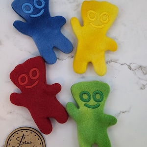 May include: Four colorful felt bear-shaped plush toys in blue, yellow, red, and green, each with embroidered facial features. The toys are arranged on a white and gray marble-patterned surface. A brown circular tag with the text "Sew Magick" is also present.