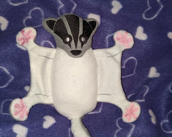 May include: White plush toy shaped like a sugar glider with a black and gray face and pink paw prints. The toy is lying on a purple blanket with white hearts.