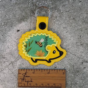 Puede incluir: Llavero de fieltro amarillo en forma de erizo con un diseño de tela verde y amarillo que presenta una abeja y flores. El llavero mide aproximadamente 5 cm de largo.