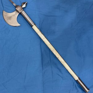Peut inclure: Une arme d'hast dotée d'une tête de hache en métal argenté poli avec une pointe supérieure acérée et un crochet arrière. Le long manche est composé d'une série de segments lisses de couleur crème insérés entre des bagues en métal argenté.