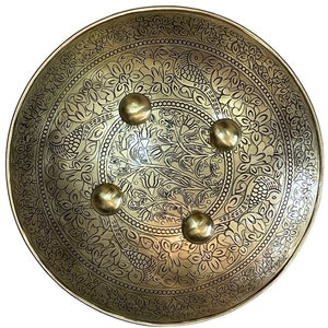 Puede incluir: Un escudo redondo de color dorado con un intrincado diseño floral y cuatro remaches de metal en relieve. El escudo está decorado con un patrón de pájaros y flores.