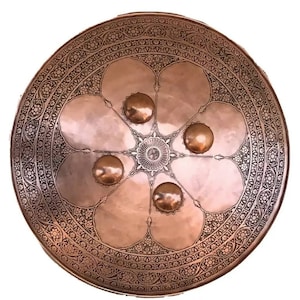 Peut inclure: Un bouclier rond en cuivre avec des motifs floraux complexes gravés à la surface. Le bouclier a cinq bossages circulaires en relief autour d'un motif central en forme d'étoile.