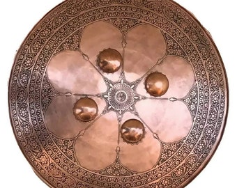 May include: A round, copper shield with intricate floral designs etched into the surface. The shield has five raised, circular bosses around a central starburst design.