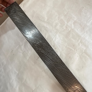 May include: A rectangular piece of steel with a unique, patterned surface. The steel has a dark, mottled appearance with flowing, wave-like designs. The metal is held by a hand, suggesting it is a raw material for crafting.