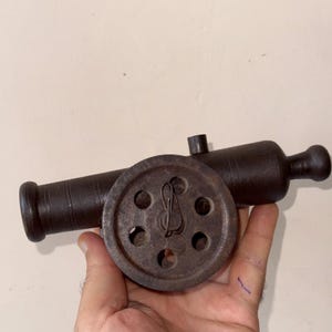 May include: A miniature, antique-style cannon made of dark metal. The cannon features a barrel, a wheel with circular cutouts, and a handle. The overall design suggests a historical or decorative item.
