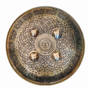 May include: A round, antique-style shield with an intricate floral and geometric pattern in gold and black. The center features a sun face design. Four silver-colored, rounded studs are evenly spaced around the center.