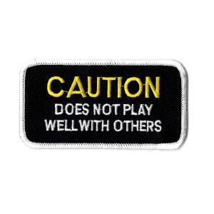 May include: Black rectangular patch with white stitched border. The patch features the word "CAUTION" in yellow block letters above the phrase "DOES NOT PLAY WELL WITH OTHERS" in white block letters.