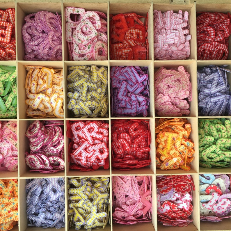 May include: A wooden box filled with colorful fabric letters. The letters are arranged in rows and columns, with each row featuring a different color and pattern. The letters are all the same size and shape, and they are made of a soft, felt-like material.