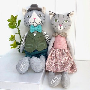May include: Two gray plush cat toys with embroidered faces. The cat on the left is wearing a green vest, a blue bow tie, and a gray hat. The cat on the right is wearing a pink dress with a floral pattern. The cat on the left has the name "Rory" embroidered on its foot. The cat on the right has the name "Katie" embroidered on its foot.