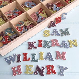 以下が含まれることがあります： カラフルな水玉模様の文字でいっぱいの木製の箱。明るい青色の表面に、SAM、KIERAN、WILLIAM、HENRYの名前が綴られています。
