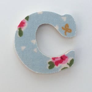 Puede incluir: Una letra C de tela azul claro con un patr&oacute;n floral. La letra est&aacute; hecha de una tela suave y texturizada con flores rosas y blancas y hojas verdes.
