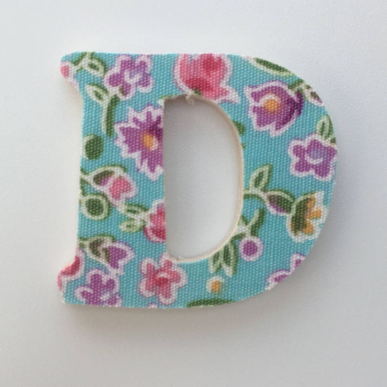 Puede incluir: Una letra D de tela azul claro con un patr&oacute;n floral. La letra est&aacute; hecha de una tela suave y texturizada con una variedad de flores rosas, moradas y verdes.