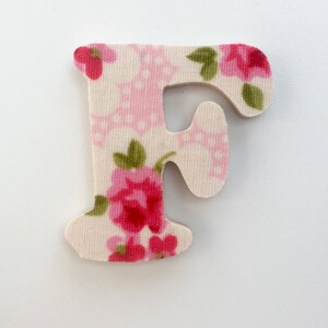 Puede incluir: Una letra F de tela blanca con un patr&oacute;n floral rosa y verde. La letra est&aacute; recortada y tiene una textura ligeramente en relieve.