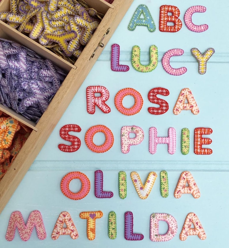 May include: A set of colorful fabric letters, including the names "ABC", "Lucy", "Rosa", "Sophie", "Olivia", and "Matilda". The letters are made of different patterned fabrics and have a stitched border.