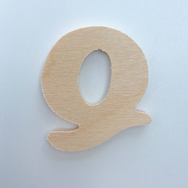 Puede incluir: Una letra Q de madera, pintada en un color madera natural. La letra est&aacute; recortada en una fuente simple y redondeada.
