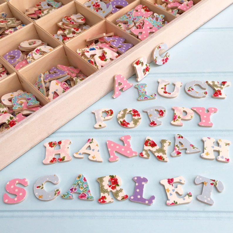 Puede incluir: Una caja de madera llena de letras de colores cubiertas de tela. Las letras est&aacute;n dispuestas para deletrear los nombres "Lucy", "Poppy", "Hannah" y "Scarlet".