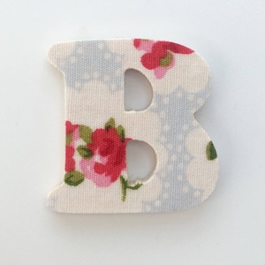 Puede incluir: Una letra B de tela blanca con un patr&oacute;n floral. La letra est&aacute; recortada y tiene un fondo blanco con flores rojas y rosas.