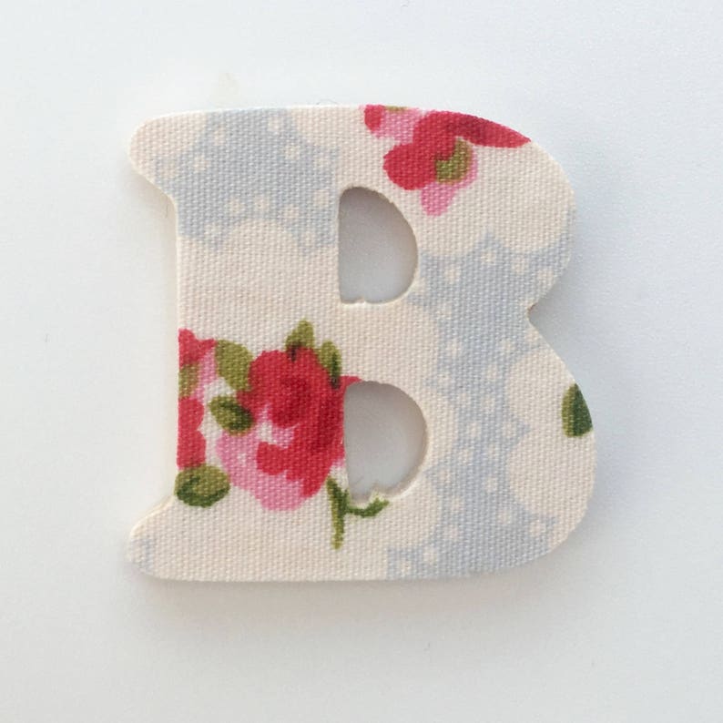 Puede incluir: Una letra B de tela blanca con un patr&oacute;n floral. La letra est&aacute; recortada y tiene un fondo blanco con flores rojas y rosas.
