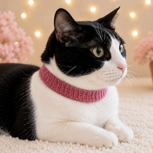 May include: A black and white cat wearing a solid dusty rose pink crocheted collar. The collar features a ribbed texture and sits snugly around the cat's neck. The cat has green eyes and is positioned in a profile view, facing toward the right side.