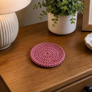 Può includere: Un sottobicchiere rotondo all'uncinetto di colore rosa antico è appoggiato su una superficie di legno. Il sottobicchiere è realizzato con un motivo a punto stretto e i bordi sono rifiniti con cura. Fa parte di un tema di arredamento per la casa.
