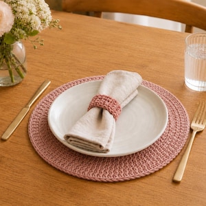Può includere: Una tovaglietta rotonda all'uncinetto color rosa polvere sotto un piatto in ceramica bianca. Un tovagliolo di lino beige è piegato e fissato con un portatovagliolo all'uncinetto abbinato. Un coltello e una forchetta dorati sono ai lati.