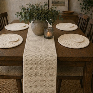 May include: A wooden dining table set with four chairs, a cream-colored crocheted table runner, and matching placemats. A vase with greenery and a candle holder are centered on the table, creating a warm and inviting dining space.
