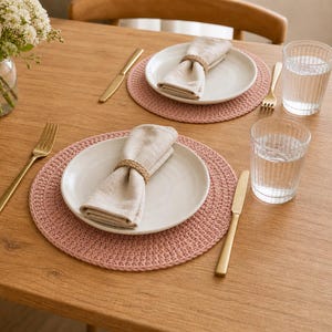 Puede incluir: Una mesa de comedor con dos cubiertos. Cada uno incluye un plato blanco, una servilleta beige doblada sujeta por un anillo de cuerda, cubiertos dorados, un salvamanteles rosa tejido y un vaso de agua. Un jarrón con flores adorna la mesa.