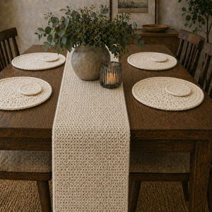 Könnte beinhalten: Ein Holztisch mit einem cremefarbenen gehäkelten Tischläufer und passenden Platzsets. Eine Vase mit Grün und ein Kerzenhalter sind auf dem Tisch zentriert, wodurch ein warmer und einladender Essbereich entsteht.