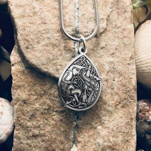 May include: A silver Celtic knot pendant necklace with a detailed design of a knot with a bird inside. The necklace is on a silver chain and is resting on a rock.