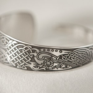 Puede incluir: Una pulsera de plata con un dise&ntilde;o de nudo celta. La pulsera es curva y tiene un patr&oacute;n detallado de nudos y espirales.