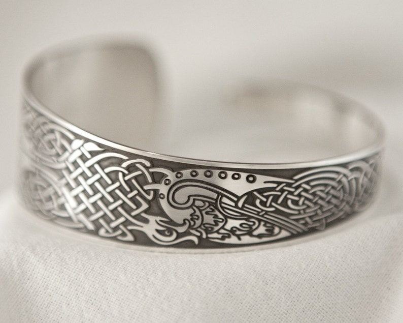 Puede incluir: Una pulsera de plata con un dise&ntilde;o de nudo celta. La pulsera es curva y tiene un patr&oacute;n detallado de nudos y espirales.