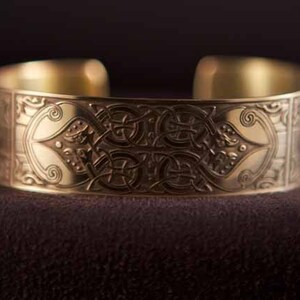 May include: A gold-toned metal bracelet with an intricate Celtic knot design. The bracelet is wide and has a smooth, polished finish.