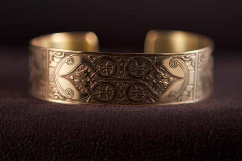May include: A gold-toned metal bracelet with an intricate Celtic knot design. The bracelet is wide and has a smooth, polished finish.