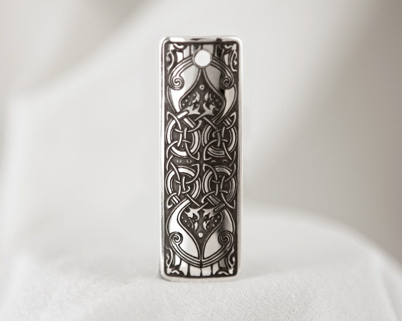 May include: A silver pendant with a detailed Celtic knot design. The pendant is rectangular and has a hole at the top for a chain.