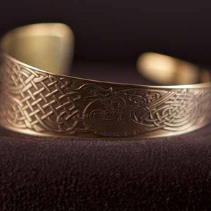 Puede incluir: Un brazalete de metal dorado con un intrincado dise&ntilde;o de nudo celta. El brazalete es curvo y tiene un acabado liso y pulido.