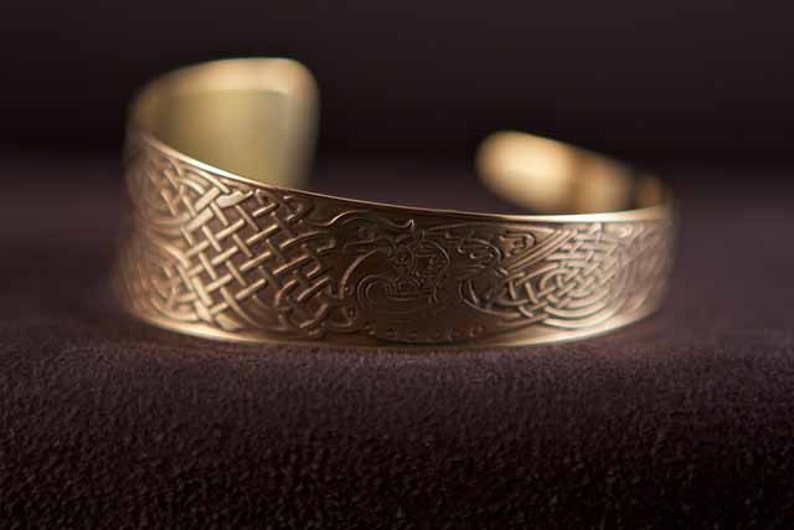 Puede incluir: Un brazalete de metal dorado con un intrincado dise&ntilde;o de nudo celta. El brazalete es curvo y tiene un acabado liso y pulido.