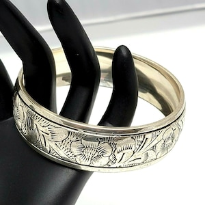 May include: A silver-toned bangle bracelet with an engraved floral pattern. The bracelet is round and wide, with a detailed design of flowers and leaves around the band. The bracelet is displayed on a black surface.