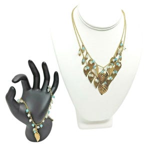 May include: A gold-toned necklace and bracelet set. The necklace features layered chains with leaf pendants and light blue and white beads. The bracelet has a similar design, displayed on a black hand-shaped jewelry holder. The necklace is displayed on a white mannequin.