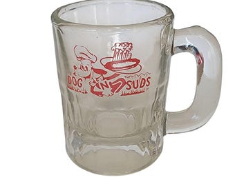 May include: Clear glass mug with a handle. The mug features a red graphic of a cartoon dog wearing a chef's hat, holding a hot dog, and the words "Dog N' Subs". The mug has a textured exterior.
