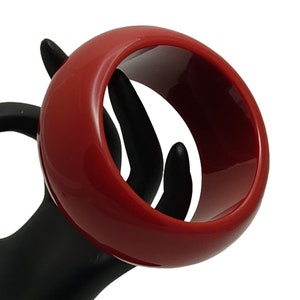 May include: A glossy, red bangle bracelet. The wide, circular bracelet is a solid color with a smooth, reflective surface. The jewelry is shown on a black display hand, creating a contrast with the red color.