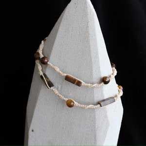 Può includere: Una collana a doppio filo con perline marroni e dorate di occhio di tigre e pietre rettangolari marroni e dorate di occhio di tigre. La collana è realizzata con un cordoncino color crema.