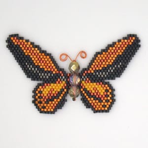 Puede incluir: Un broche de mariposa con cuentas de color naranja, negro e iridiscente. La mariposa tiene antenas de alambre.
