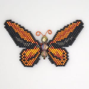 Puede incluir: Un broche de mariposa con cuentas de color naranja, negro e iridiscente. La mariposa tiene antenas de alambre.