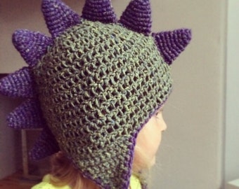 May include: A purple and green crocheted hat with a dinosaur design. The hat has a long tail and spikes on top.