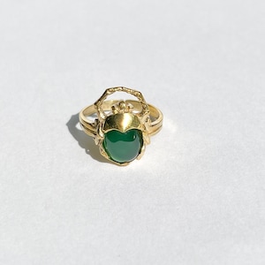 May include: A gold-colored ring featuring a scarab beetle design. The ring has a large, oval-shaped, green gemstone at its center. The beetle's details are visible, with a textured body and intricate legs. The ring is set against a white background.