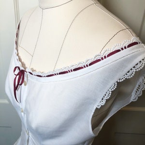 May include: White cotton blouse with a scoop neckline, adorned with delicate white lace trim and a burgundy ribbon detail. The blouse features small, round buttons down the front and is displayed on a mannequin.